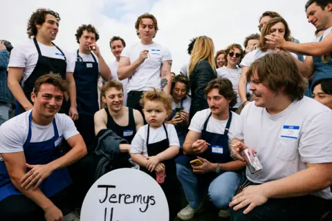 AP A crowd of Jeremy Allen White lookalikes, including a toddler, dressed in chef's aprons and white t-shirts, crouch and stand in a half-circle in a park in Chicago
