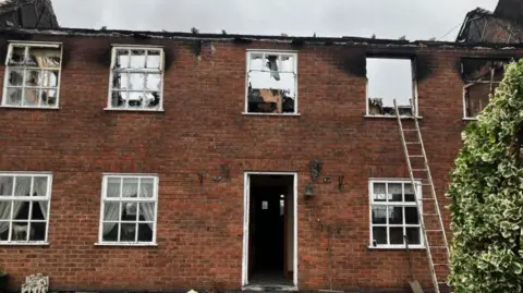 An outside view of the aftermath of the house fire with a ladder leading up to the top windows which are completely gutted with no roof