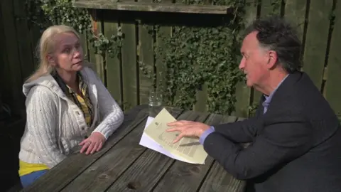 Tina Nash wearing a white cardigan and a yellow gingham shirt sits at a park bench with MP Andrew George who is wearing a grey suit. He is holding some pieces of paper in his hands. A fence to the side of them has ivy growing on it.