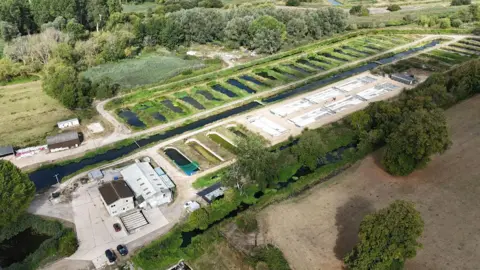 Julian Connor A drone shot of the site, with a series of ponds surrounded with greenery going down one side of a large channel which lookts like a river but isn't. On the other side, a couple of smaller industrial buildings and around six concrete tanks.