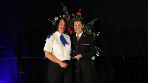 Dyfed-Powys Police Rachel standing alongside a sergeant who is holding her trophy and has her arm around Rachel's shoulder. They are both stood on a dark stage with a large flower display behind them. Both women are looking and smiling at the camera. Rachel, on the left, is wearing a white shirt with a blue tie and black trousers. The officer next to her is wearing a black suit, a white shirt and a black and white chequered tie.