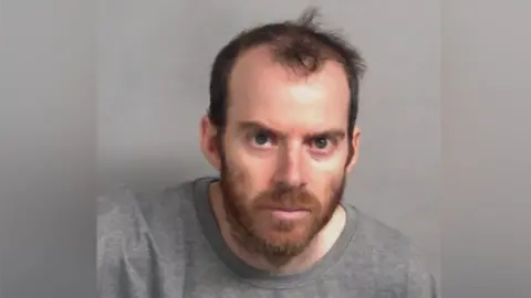 A custody shot of a man looking directly at the camera while wearing a grey jumper. He has a brown beard and hair. 