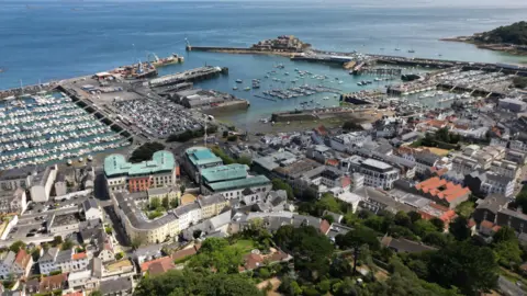 An aerial view of Guernsey on a sunny day. It shows the harbour and harbour wall in St Peter Port, with Castel Cornet on the far side.