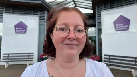 Heather Cliffe is outside a building at Bishop Grosseteste University. She has round gold glasses and plum coloured hair shaped into a bob.