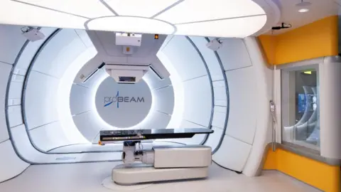 UCLH A futuristic-looking hospital room. There is a white circular backdrop, a shiny black surface to lie on, and instruments protruding from the ceiling above.