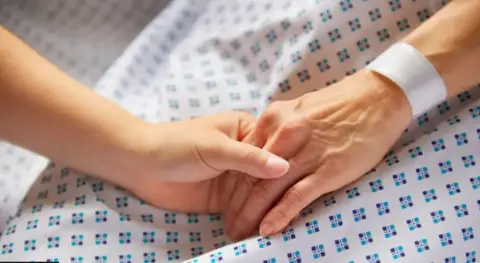 Two people hold hands on a medical bed. A younger hand clasps that of an older adult.