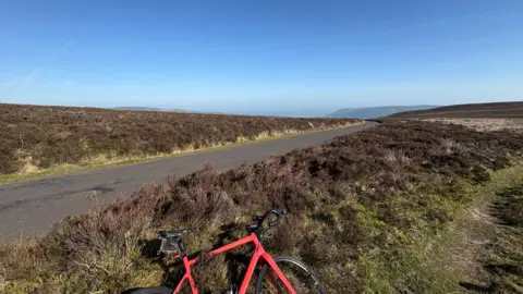 Mike Bradley A photo of Dunkery Beacon in Exmoor, Somerset. Rolling moorland under a blue sky. A single-lane road cuts through the picture and a red bike lies in the foreground.
