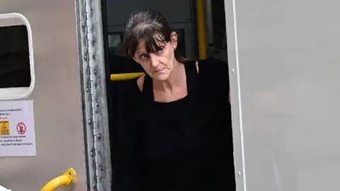 A woman with dark hair walking out of a police van. She had a black jumper dress on