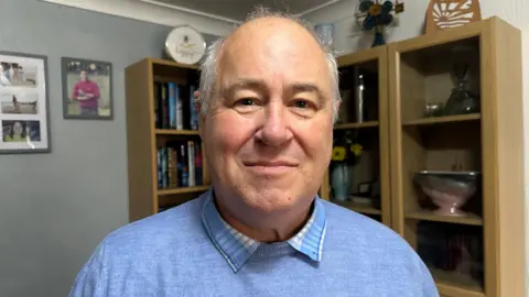 Steve Hodgson, wearing a blue jumper and shirt and standing in front of a cabinet at his home in Telford, with photos in frames on the wall behind him.