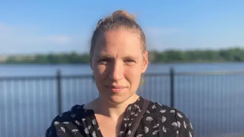 A woman with blond hair, tied back, a black top and a neutral expression. She is standing in front of a metal railing in front of a river. In the background, on the other side of the river, we can see trees and greenery. It is day time and a blue sky can be seen.