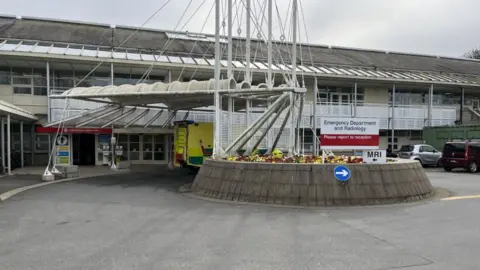 BBC Princess Elizabeth Hospital, La Vauquiedor, St Martin, Guernsey. The image shows the entrance to the Emergency Department and Radiology unit. There is a grey concrete roundabout in front of the entrance with an ambulance parked half out of sight behind the roundabout. The building is grey and cream in colour and comprised of two storeys.