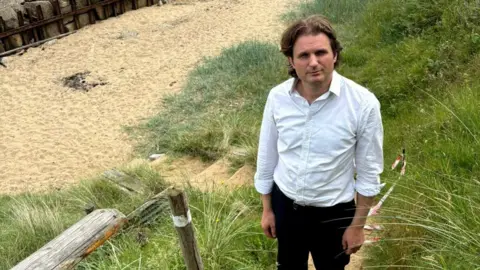 Steffan Aquarone A man wearing a white shirt and dark trousers looks towards a camera while standing on some steps that lead down to a beach. 