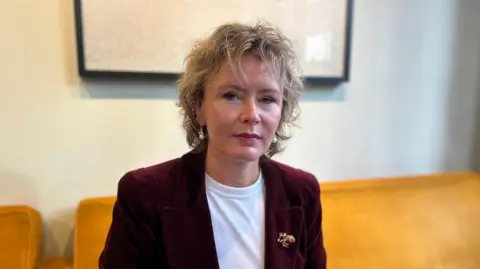 Alisa is sitting on an orange couch in an office. She is wearing a white t-shirt and a maroon velvet jacket.
