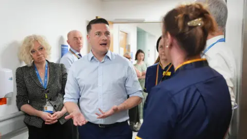 PA Wes Streeting, in a blue shirt, stands in a hospital corridor speaking to staff. 