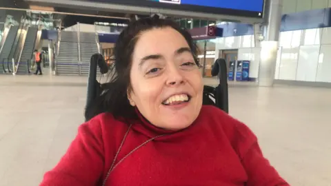 BBC Joanne Sansome - dark brown haired woman wearing a red jumper and sitting in a wheelchair at Belfast's Grand Central Station