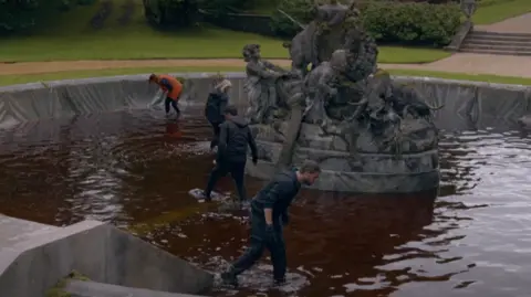 BBC/Studio Lambert A picture of contestants in the Traitors in a pool of water, with a fountain behind them.