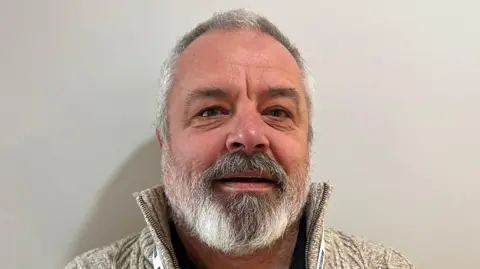 Norman Hagan has short hair and a beard. He is wearing a wooly white fleece. 