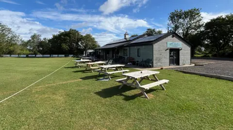 Luke Deal/BBC A landscape image showing the Battisford and District Cricket Club pavilion and its large green cricket pitch. There are several picnic tables behind the boundary rope.