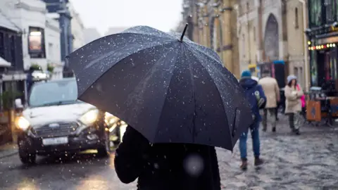 Getty Images A man walks down a road in Brighton city centre, his back to the camera, carrying an umbrella in heavy rain. Other passers by and a car can be seen outside the entrance to the Brighton Pavilion.