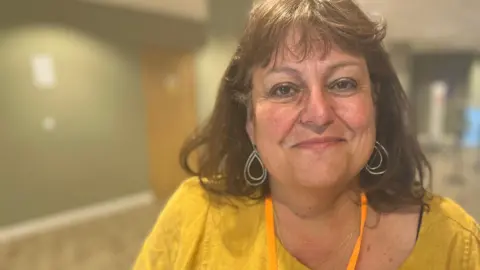 Head and shoulders image of Sarah Conboy in a hallway. She has shoulder length brunette hair, is wearing rounded earrings and a yellow top.