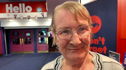 John Devine/BBC Margaret Heaney has sandy coloured thinning hair parted to the right, she has gold-coloured glasses and is wearing a beige woollen top with black lines around the collar and shoulders, she is in the foyer of a bingo hall with two set of purple double doors behind her and a blue carpet.