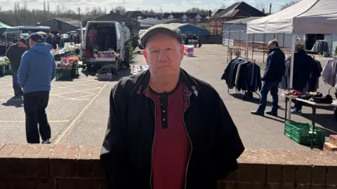 LDRS Geofrey Cripps is wearing a dark coat over a red jumper. He also has a cap on. Behind him, market stalls are set up on a car park.