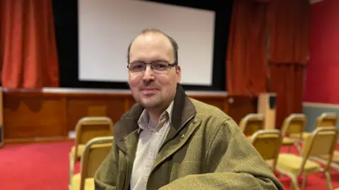A man with short, balding hair and glasses wearing a green tweed jacket and a neutral shirt. He is sitting on a chair, but has turned to face the camera, which is behind him. There is a large screen and red cinema curtains in the background.