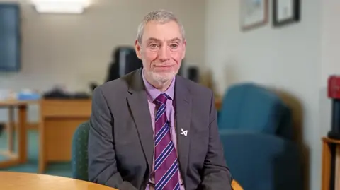 Hugh Hume. He has short grey hair, wearing a dark grey blazer with a silver sin on the lapel, light purple shirt and purple striped tie. He is sitting on a chair beside a table in an office.
