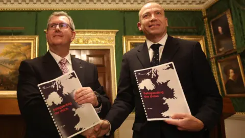 Pacemaker Jeffrey Donaldson and Chris Heaton-Harris shake hands and hold up a ring-bound paper entitled Safeguarding the Union. Both are in suits and are in a very formal-looking room with paintings in gold frames.