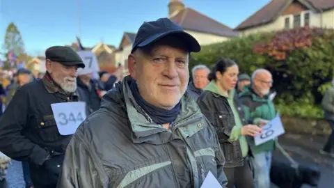 A man in a cap and a dark-coloured jacket. He is looking at the camera and is stood in a road surrounded by several other people.