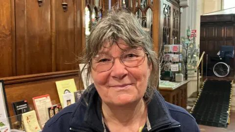 Jane has large glasses and messy grey hear. She is smiling and wearing a navy double-collared fleece. Behind her are religious pamphlets arranged along a shelf. 