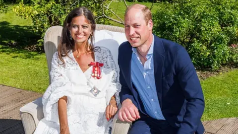 Princew William in a blue suit and shirt crouched next to Dame Deborah James, in a white dress with a medal pinned to it.