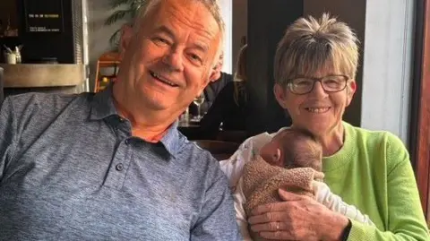 NHS Blood and Transplant handout Left to right: Tad with grey hair wearing a grey polo top sitting alongside Nicki Detko with short grey and blond hair holding one of her grandchildren who is sleeping wrapped in a blanket. Nicki is wearing a lime green jumper. They are sitting in a restaurant smiling.