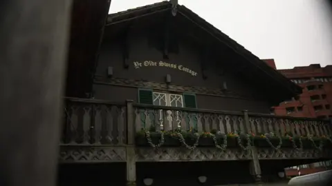 The upper facade of Ye Olde Swiss Cottage pub, show its dark chalet-style architecture with wooden balconies, decorative railings, and window shutters. A row of colorful flowers adorns the balcony, while modern red-brick buildings rise in the background under an overcast sky.