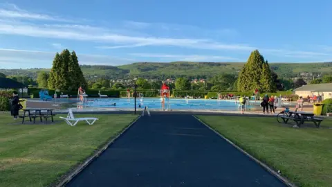 An outdoor swimming pool surrounded by hills and trees. At the side of the pool are a number of picnic benches and people