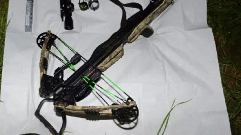 Hertfordshire Police A crossbow, which appears to have a camouflage-like trim and black metallic holder, lying on a white sheet on top of grass