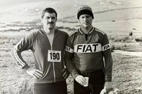 Bill Anderson A black and white picture of Mr Allan and Mr Anderson in running clothes surrounded by hills. Mr Anderson is wearing a woolly hat. Mr Allan has a moustache.