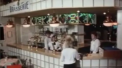 A blurry image shows a smart looking brasserie bar with white tiles on the front. There are five waiters wearing white shirts and black trousers or skirts. There are pendant lights hanging down.