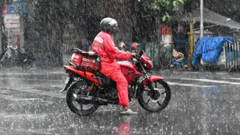 A food delivery person from Zomato - wearing a red uniform and helmet - is waiting at a traffic signal on his bike, during heavy rain in Kolkata, India. 