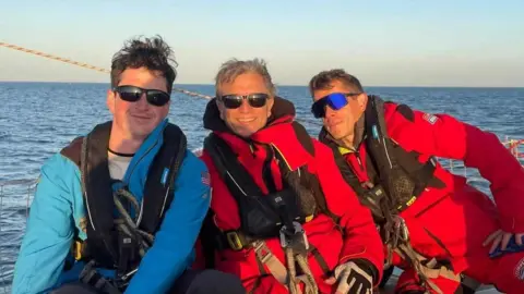 Kevin Lynch Kevin Lynch is far right of this three man photo. He is wearing a red full body sailing suit, with life-jacket and has sunglasses on. There is the ocean in the background.