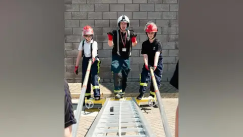 Northamptonshire Fire and Rescue Service An instructor wearing firefighting protective equipment stands behind two participants in helmets and gloves as they practice extending and handling a ladder during a training activity beside a brick wall.