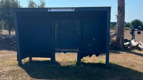 A dark blue sports team shelter stands on a dry grass field. The shelter has a damaged side panel with a large hole. Cut tree trunks, stacked logs, and a lamp post are visible in the background under a clear blue sky.