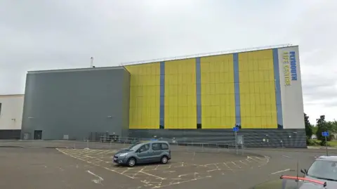 Google Maps Plymouth Life Centre building exterior. The building is green and grey. A car park is in front of the building. A grey car is parked in a disabled bay.