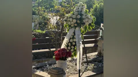 Callum Dickson A grave site has red flowers in an urn and a cross-shaped floral tribute made of white roses. It is placed on a stand on top of the grave. Trees can be seen in the background.
