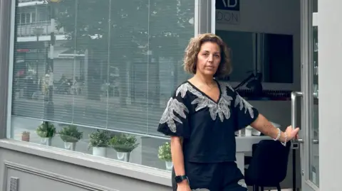 Eleni Lasheen outside her salon. She has wavy dark-blonde hair and is wearing a black and silver patterned top
