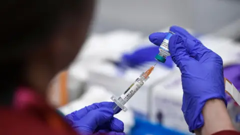 Close up of a nurse wearing blue latex gloves extracting a vaccine from a small glass bottle with a syringe. 