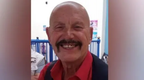 West Yorkshire Police Kurt Giwisser wearing a bright red collared shirt underneath a dark vest. The shirt is buttoned down the front, and the vest appears to be sleeveless. The background includes a blue railing and a light-coloured wall. There are some posters visible on the wall behind him, and another individual is partially visible to the left side of the image, seated and wearing a light-coloured top.