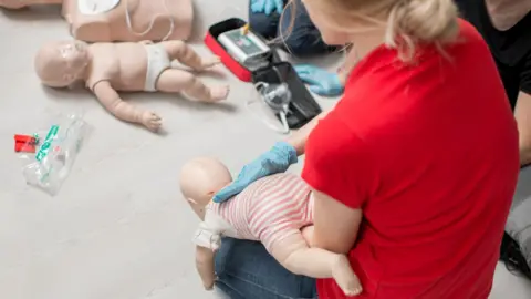 Getty Images A first aid training session showing what to do if a small child is choking.  A trainer has her back to the camera and is kneeling down, holding a mannequin baby face-down on her thigh. She has blonde hair and is wearing a red t-shirt and blue jeans. Two other mannequins - a baby and an adult - are placed on the floor in front of the trainer. 