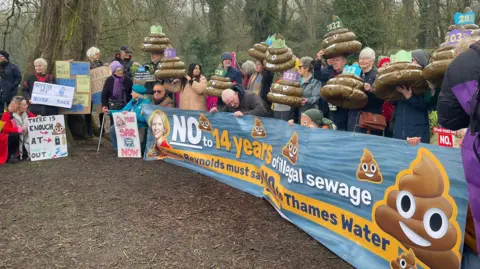 We Own It A group of people in a line on the bank of a river. They are holding a long banner with a picture of a poo emoji and the words "No to 14 years of illegal sewage", and several smaller banners. Some have the emojis on their heads.