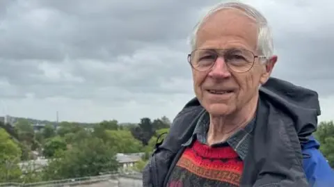 Andrew Lack has combed over white hair and is wearing a coat over a jumper.